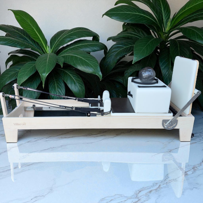 Full Side View of Milaron Maple Studio Reformer with Creme Carriage and Frame with accessories sitting on carriage and jumpboard attached with greenery plants in background
