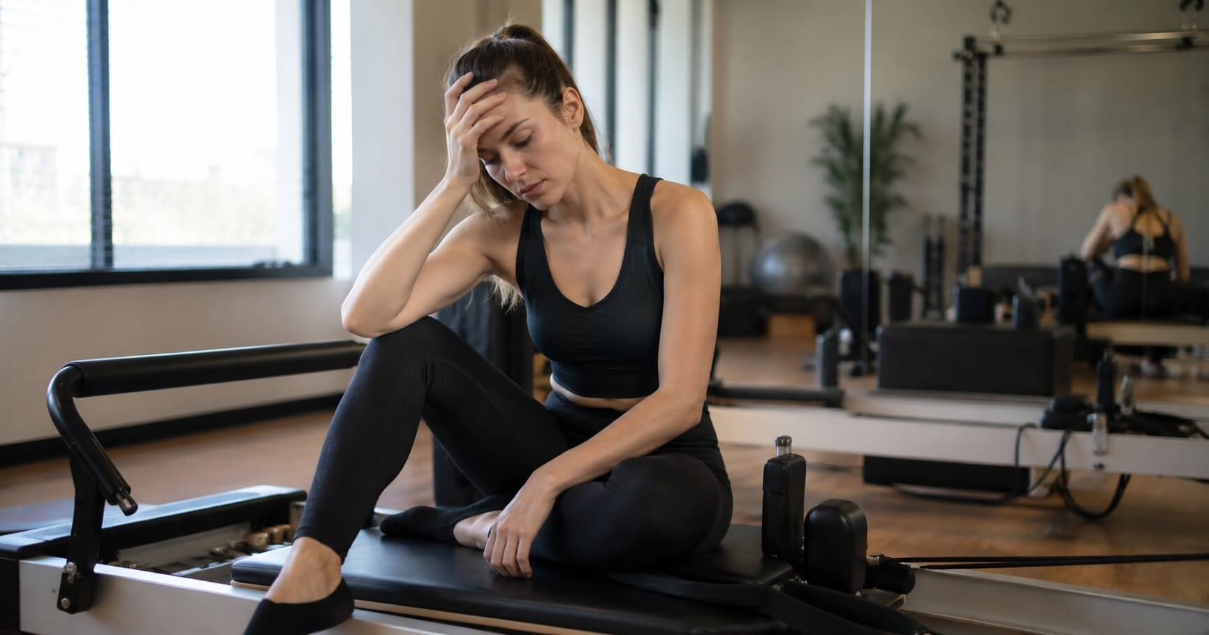 A woman in black activewear sits on a Pilates reformer machine in a bright fitness studio, holding her head in her hand in a moment of exhaustion or discomfort