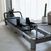 Full View of Queen Foldable Reformer from front with reformer in open position showing white frame and grey upholstery in home studio setting