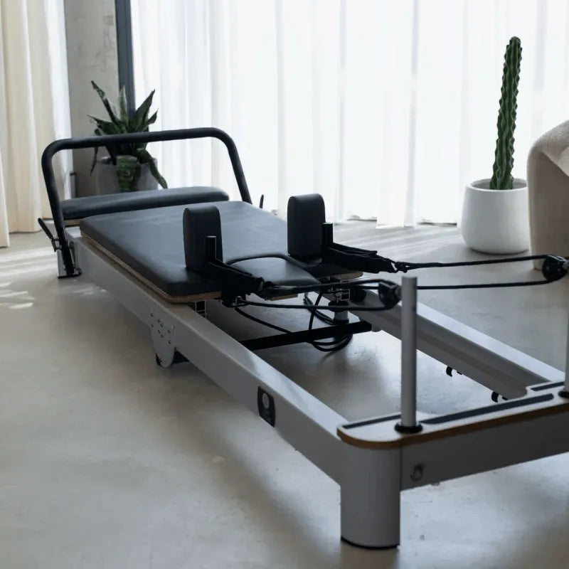 Full View of Queen Foldable Reformer from front with reformer in open position showing white frame and grey upholstery in home studio setting