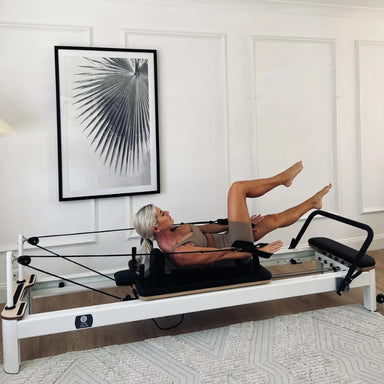 Core Collab Queen Reformer white frame with black upholstery in home setting with lady on top doing exercises