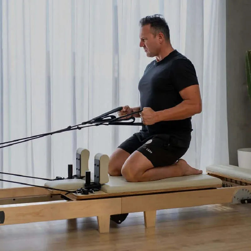 Man using a Pilates reformer machine in a home setting kneeling and using arm straps