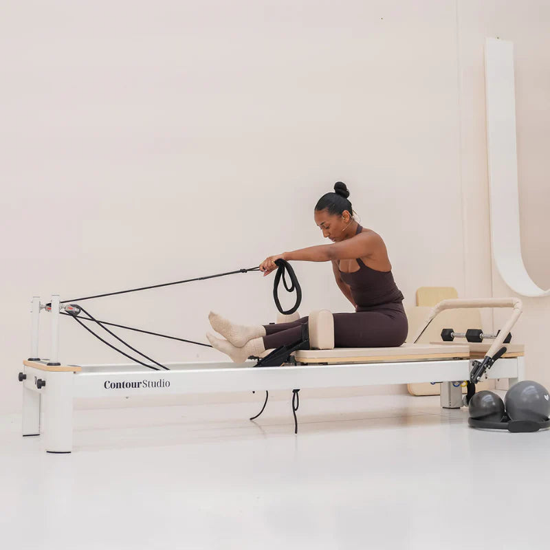 Side view of lady sitting on white pilates reformer leaning forward with hands in straps