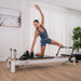 Woman in side pose and foot on footbar of black and white pilates reformer in home setting