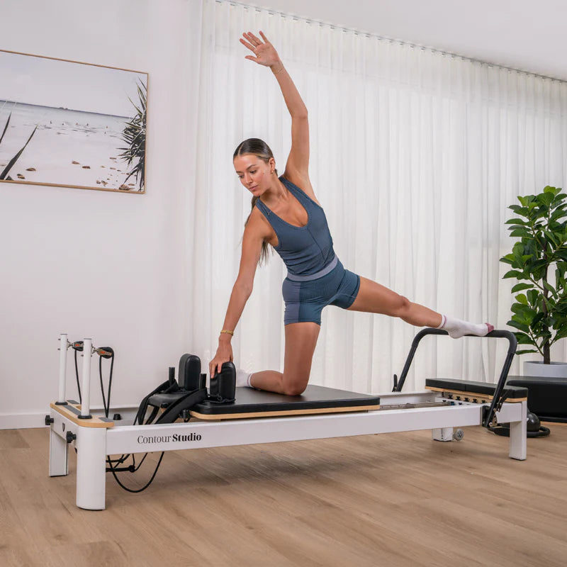 Woman in side pose and foot on footbar of black and white pilates reformer in home setting