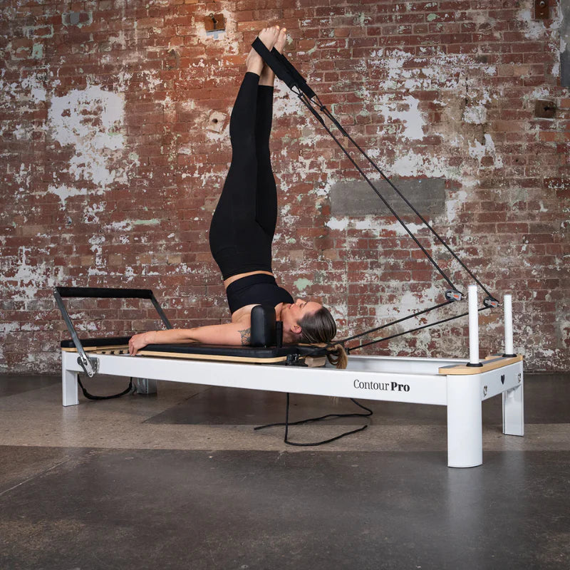 Person using a  White Aluminium Pilates reformer machine against a brick wall.