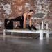 Woman exercising on a  White Aluminium Pilates reformer machine against a brick wall.