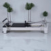 Milaron Blanc Pilates reformer machine showing full side view with white frame and black carriage in white room with plants in background