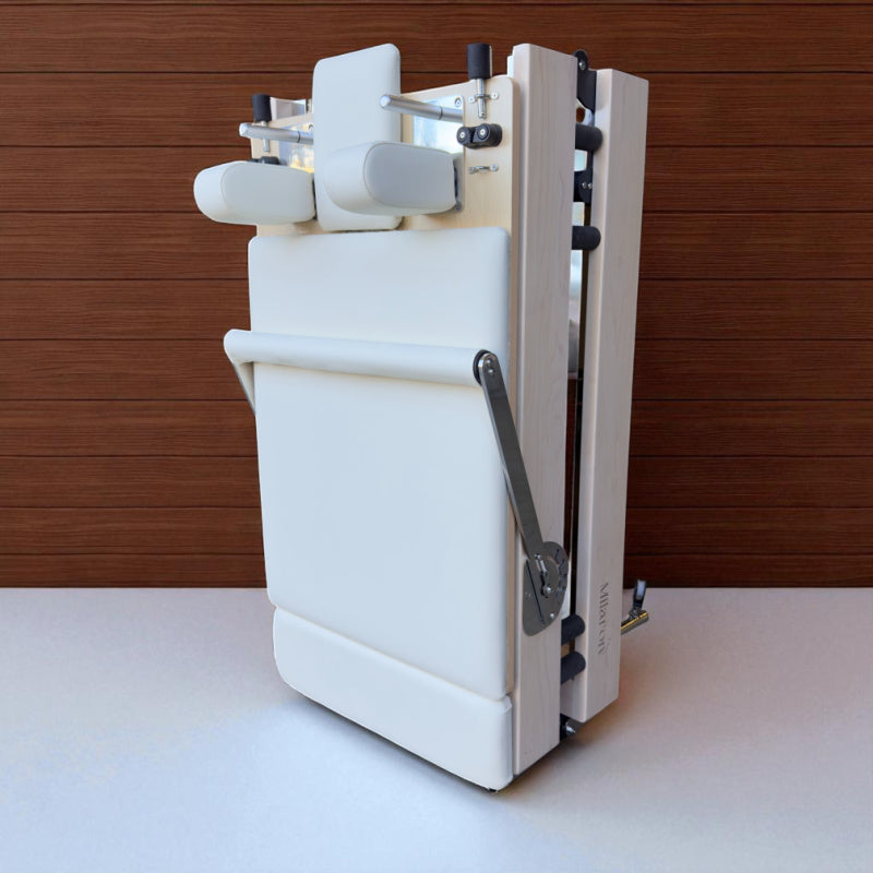 Angled view of Milaron Maple foldable reformer in folded position shown in creme upholstery and standing upright
