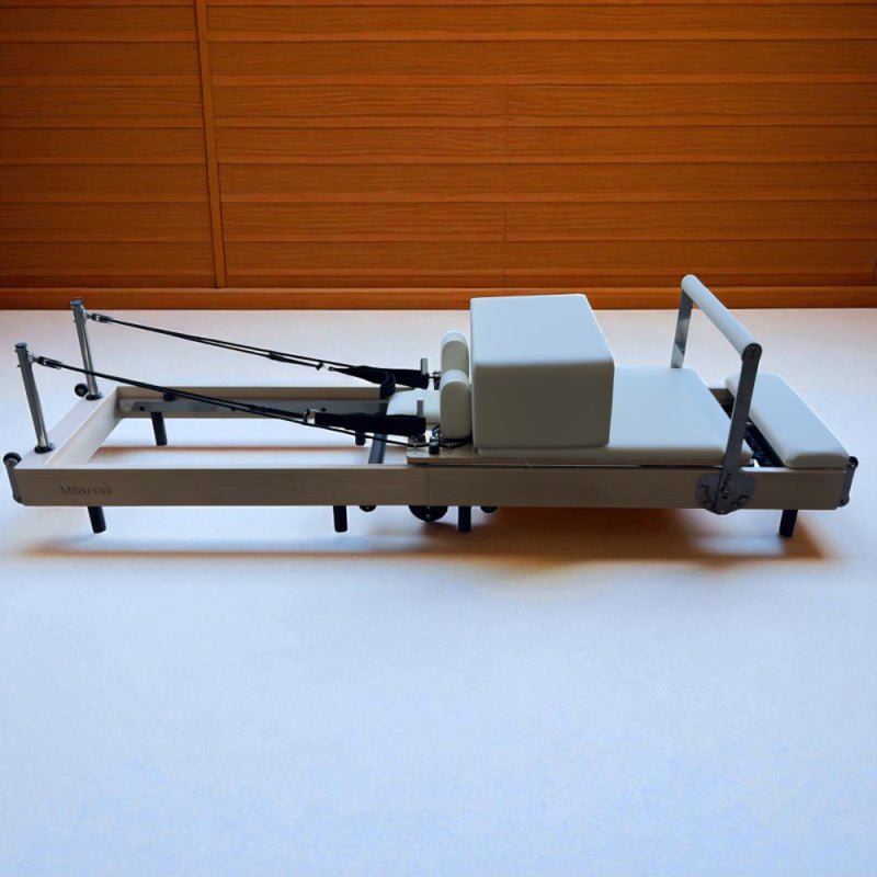 Full side view of Milaron Maple Foldable reformer in studio setting with straps hooked on to hooks near headrest. Also shows sitting box sitting on the carriage