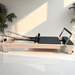 Milaron Pilates Reformer with Maple Wood Frame and Black Upholstery indoors on white floor with indoor plants