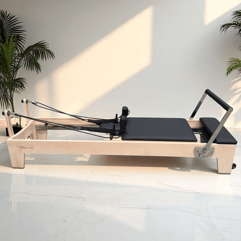 Milaron Pilates Reformer with Maple Wood Frame and Black Upholstery indoors on white floor with indoor plants