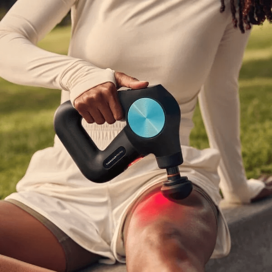 Seated female using Theragun PRO Plus massage gun with infra red light to massage quads
