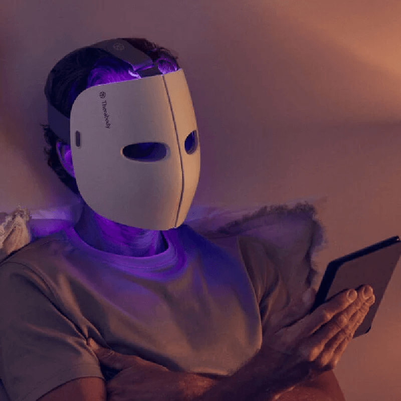 Male sitting in bed reading kindle with white Theraface Mask on with blue LED lights